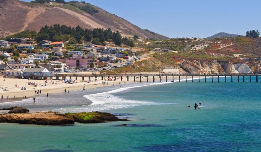 Aerial coastal view of a sandy beach with a long pier, turquoise ocean water, and hillside homes