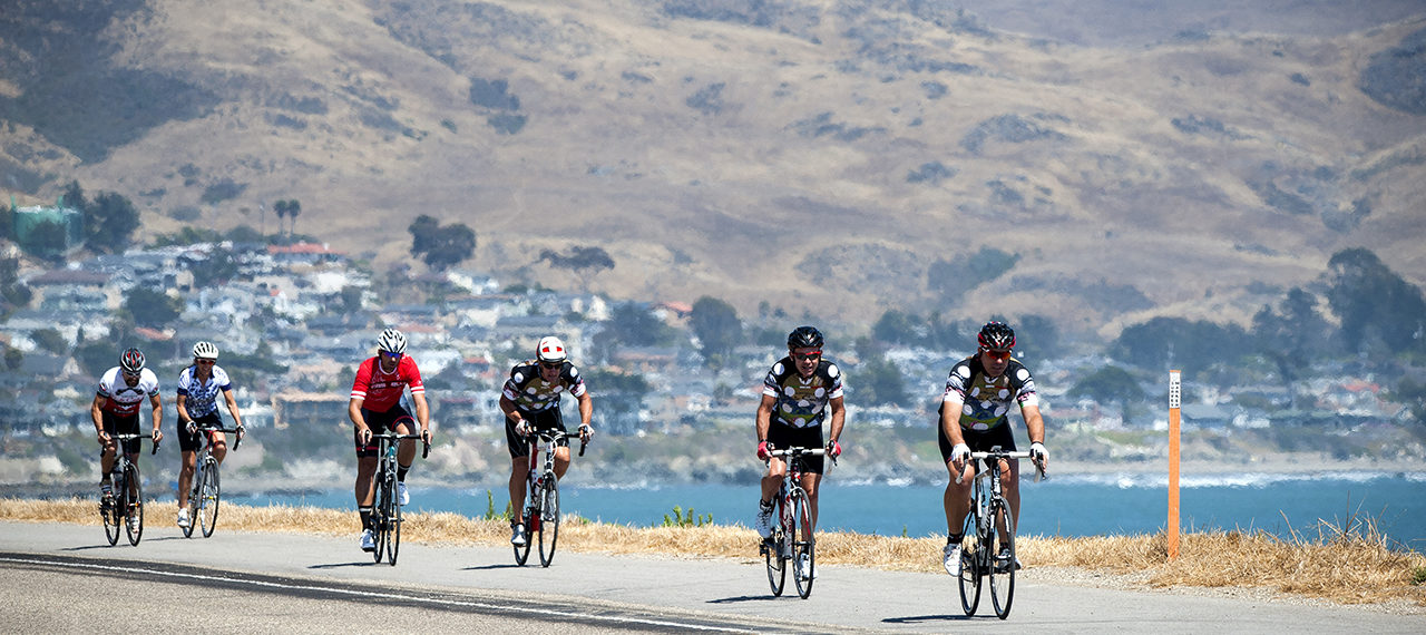 Cyclists riding along a coastal road with hills and a town visible in the background in bright daytime light